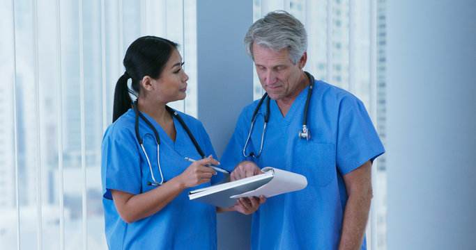 Wall Mural -  - Medium shot of two friendly medical professionals wearing blue scrubs working together. Japanese woman doctor and Caucasian male nurse going over paperwork in hospital