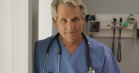 Wall Mural - Close up portrait of senior Caucasian male doctor or nurse looking into camera lens while standing by window in hospital. Older man medical professional wearing blue scrubs