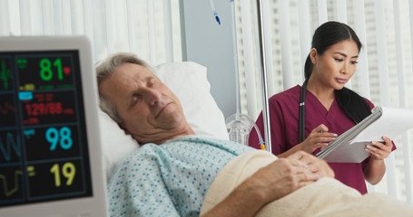 Wall Mural - Doctor or nurse taking notes on chart with senior Caucasian male patient recovering in hospital bed in foreground. Japanese woman medical professional monitoring recovery