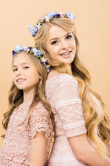 Wall Mural - attractive mother and adorable child in colorful floral wreaths standing back to back and looking at camera on yellow background