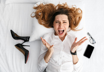 Poster - Displeased young business woman dressed in formal clothes shirt indoors lies on bed near shoes and phone.