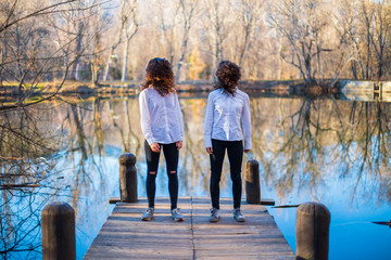 Young twin sisters in same clothes shaking hair at lake on autumn day in the forest