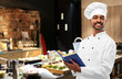 © Syda Productions - cooking, profession and people concept - happy male indian chef in toque with cookbook over restaurant kitchen background