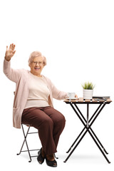 Sticker - Senior woman sitting at a table with a cup of coffee and greeting with hand