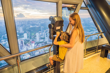 Wall Mural - Mom and son are looking at Kuala lumpur cityscape. Use binoculars. Panoramic view of Kuala Lumpur city skyline evening at sunset skyscrapers building in Malaysia. Traveling with kids concept