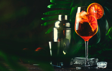 Aperol spritz cocktail in big wine glass with bloody oranges, summer Italian fresh alcohol cold drink. Wooden bar counter background with tools, summer mood concept with palm trees, selective focus