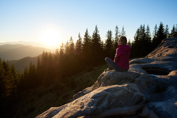 Young woman hiker sitting on big rock on the top of mountain in the evening. Female tourist enjoying incredibly beautiful sunset. On background forest, setting sun and blue sky