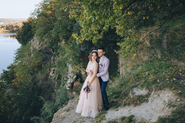  Autumn portrait of newlyweds in love, against a background of green trees, rocks and hills. Wedding photography. Stylish groom hugs a beautiful bride in a beige dress with a crown.