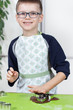 Uśmiechnięty szczerbaty chłopiec w okularach i fartuchu kuchennym wyciska kształty z metalowych foremek w brązowym cieście.