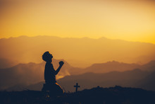Young Man Kneeling Down And Praying With Christian Cross At Sunset Background. Christian Silhouette Concept.