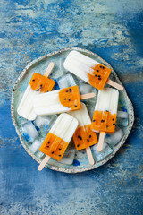 Poster - Mango passion fruit popsicles with coconut cream on ice cubes over blue plate. Healthy vegetarian vegan tropical summer snack. Copy space, blue stone background