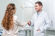 © LIGHTFIELD STUDIOS - cheerful dentist standing with hand in pocket and shaking hands with female patient