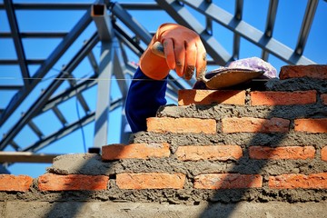 Sunlight and shadow on surface of bricklayer's hand in rubber glove using trowel to build brick wall and blurred roof structure in blue sky background