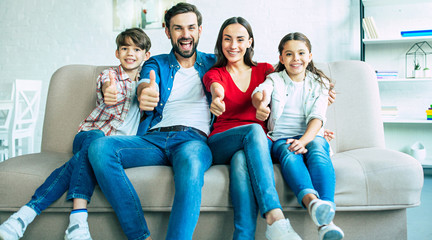 Wall Mural - We are great. Smiling beautiful modern family is happy and show thumbs up on camera while sitting on the couch