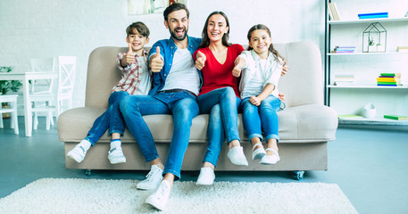 Wall Mural - We are great. Smiling beautiful modern family is happy and show thumbs up on camera while sitting on the couch