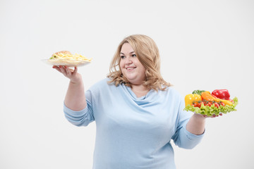 Young lush fat woman in casual blue clothes on a white background holding a vegetable salad and a plate of fast food, hamburger and fries. Diet and proper nutrition.