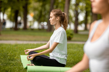 Wall Mural - Young slim girl sits in the lotus position with closing eyes doing exercises with other girls on green grass in the park on a warm day.