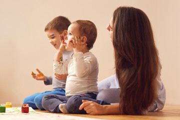 Young mother and her two little sons dressed in home clothes are sitting on the wooden floor in the room and painting with fingers on the white paper