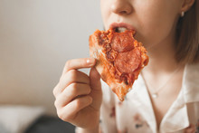 Girl In A Pajama Bites A Delicious Pizza Piece, Close-up And Enjoyment. Woman Eating Pizza, Cropped Photo.Background. Copyspace