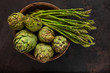 Close up photo of fresh artichoke in the old wooden bowl and Bunch of green asparagus. Top view on dark background.