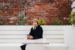 © Cavan Images - Portrait of senior woman wearing warm clothing using smart phone while sitting on bench against brick wall in city