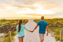 Happy Young Couple In Love Walking On Romantic Evening Beach Stroll At Sunset. Lovers Holding Hands On Summer Holidays In Florida Beach Vacation Destination. People Walking From Behind.