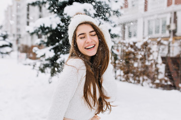 Wall Mural - Happy winter moments of joyful young woman with long brunette hair, white winter clothes having fun on street in snowing time. Expressing positivity, true brightful emotions, smiling with closed eyes