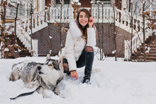 White Girl With Amazing Smile Posing With Her Dog During Winter Walk In Yard. Outdoor Photo Of Cheerful Lady Wears Ripped Denim Pants Sitting On The Snow With Lazy Husky.