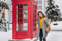 Smiling Girl In Elegant Attire Talking On Smartphone While Standing Near British Phone Booth In Winter. Outdoor Photo Of Pleased Brunette Woman In Trendy Coat Carrying Brown Backpack During Walk.