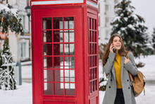 Refined Caucasian Female Model Laughing Near Red Phone Booth In Winter Morning. Outdoor Portrait Of Graceful Girl In Yellow Sweater And Coat Talking On Smartphone With Snowy Trees On Background.