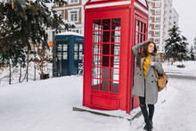 Full-length Portrait Of Magnificent Young Woman In Stylish Coat Posing With Pleasure Near British Phone Booth. Photo Of Gorgeous Dark-haired Female Model Enjoying Photoshoot In Winter Park..