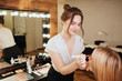 © estradaanton - Young woman putting make up and eyeshadows on face of blonde model. She stand in front of her in beauty room. Accurate and concentrated.