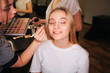 © estradaanton - Nice young woman sit in beauty room with closed eyes. She smile. Make up artist put eyeshadows on eyeline. She hold palette in hands. Another profesional stand behind.