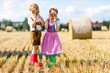 © Irina Schmidt - Two kids in traditional Bavarian costumes in wheat field. German children eating bread and pretzel during Oktoberfest. Boy and girl play at hay bales during summer harvest time in Germany.