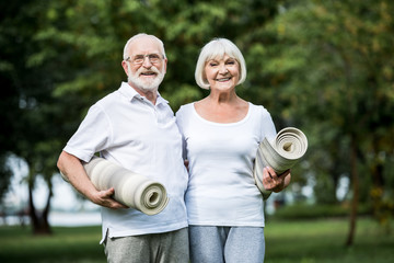 smiling senior couple holding fitness mats while standing in park