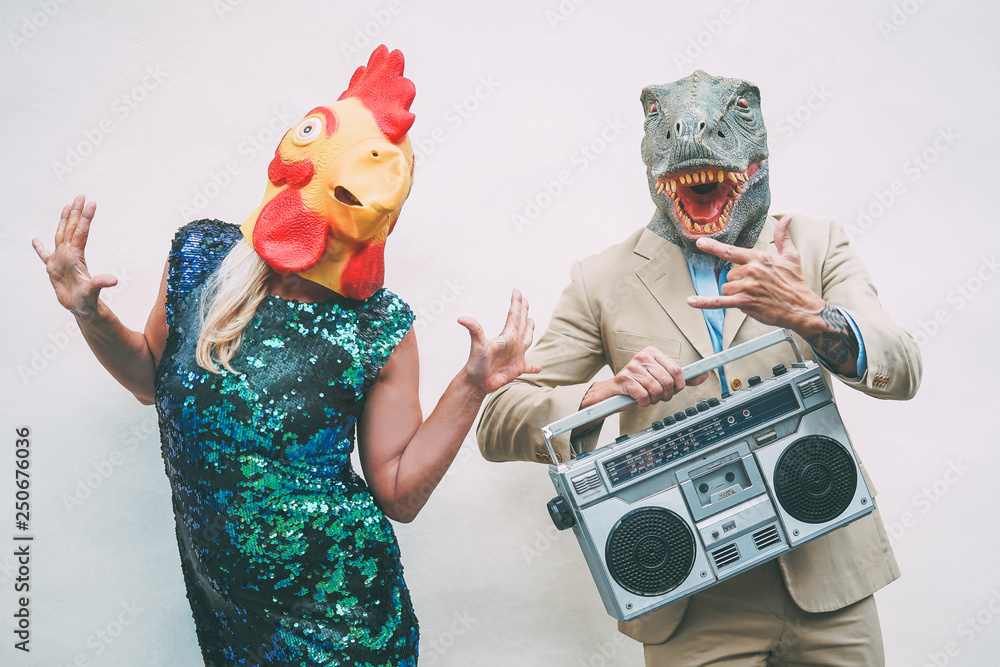 Crazy senior couple wearing chicken and t-rex mask while dancing outdoor - Mature trendy people having fun celebrating and listening music with boombox - Absurd concept of masquerade funny holidays - obrazy, fototapety, plakaty 