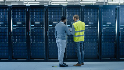 Poster - Bearded IT Specialist in Glasses and High Visibility Vest with a Laptop Computer and Black Technician Colleague Talking in Data Center while Standing Next to Server Racks. Running Maintenance. 