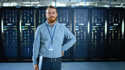 Poster - Bearded IT Specialist in Glasses is Standing in Data Center Next to Server Racks, Smiling to the Camera and Holding Laptop. Running Diagnostics or Doing Maintenance Work.