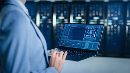 Back View Close Up Shot of an IT Specialist Working on Laptop in Data Center Next to Server Racks. Computer is Running Diagnostics or Doing Maintenance Work.