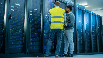 Aufkleber - IT Administrator with a Laptop Computer in High Visibility Vest and Black Male Engineer are Talking in Data Center while Standing Before a Server Rack. Running Diagnostics or Doing Maintenance Work. 