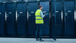 © Gorodenkoff - IT Engineer in High Visibility Vest is Working on Laptop in Data Center while Standing Before Server Rack. Running Diagnostics or Doing Maintenance Work.
