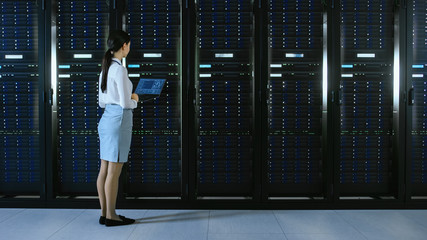 Leinwandbilder - Beautiful Data Center Female IT Technician Walking Through Server Rack Corridor with a Laptop Computer. She Stops and Visually Inspects Working Server Cabinets.