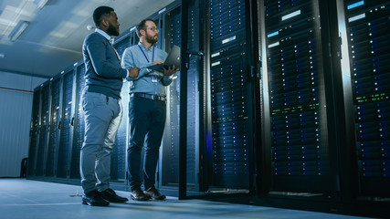 Bearded IT Technician in Glasses with a Laptop Computer and Black Male Engineer Colleague are Talking in Data Center while Working Next to Server Racks. Running Diagnostics or Doing Maintenance Work. 