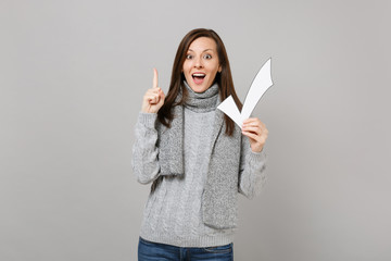 Excited young woman in gray sweater scarf holding index finger up with great new idea, hold paper check mark isolated on grey background. Healthy fashion lifestyle people emotions cold season concept.