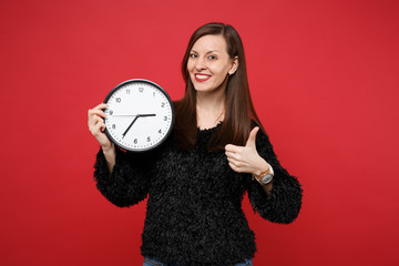 Portrait of stunning young woman in black fur sweater showing thumb up, holding round clock isolated on bright red background in studio. People sincere emotions, lifestyle concept. Mock up copy space.