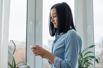 Wall Mural - Portrait of young smiling woman in shirt at home near the window with smartphone reading text