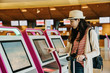 © PR Image Factory - young asian female business traveler using self service check in machine at airport lobby indoor. girl photographer carrying camera finger touching screen choosing picking on auto counter concept.