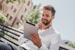 © Friends Stock - Social life. Young smiling man sitting on a bench in the park, holding his tablet. Rest, communication and relax concept