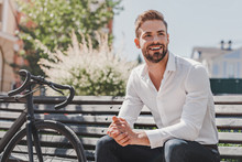 Ready To Go. Young Smiling Man Sitting On A Bench In The Park With A Bicycle Beside Him. Rest And Relax Concept