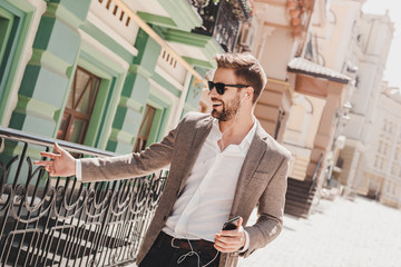 Success is how high you bounce when you hit bottom. Smiling brown-haired man in sunglasses standing outdoors with his smartphone. He holds his phone with earphones and touches fence beside him